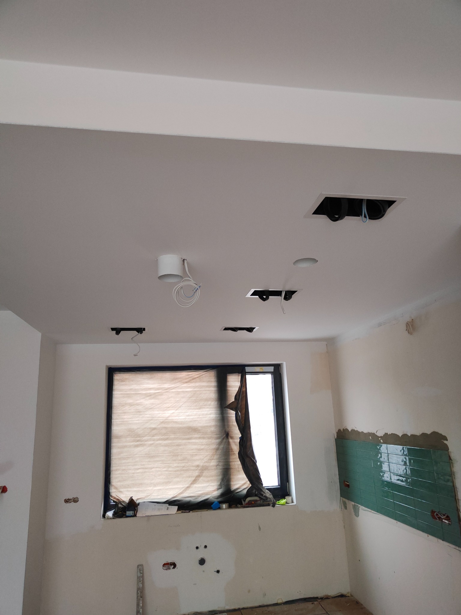 Wnętrze w trakcie remontu: widoczne przygotowane otwory w suficie pod oświetlenie, częściowo położone zielone płytki na ścianie, okno zabezpieczone folią malarską.