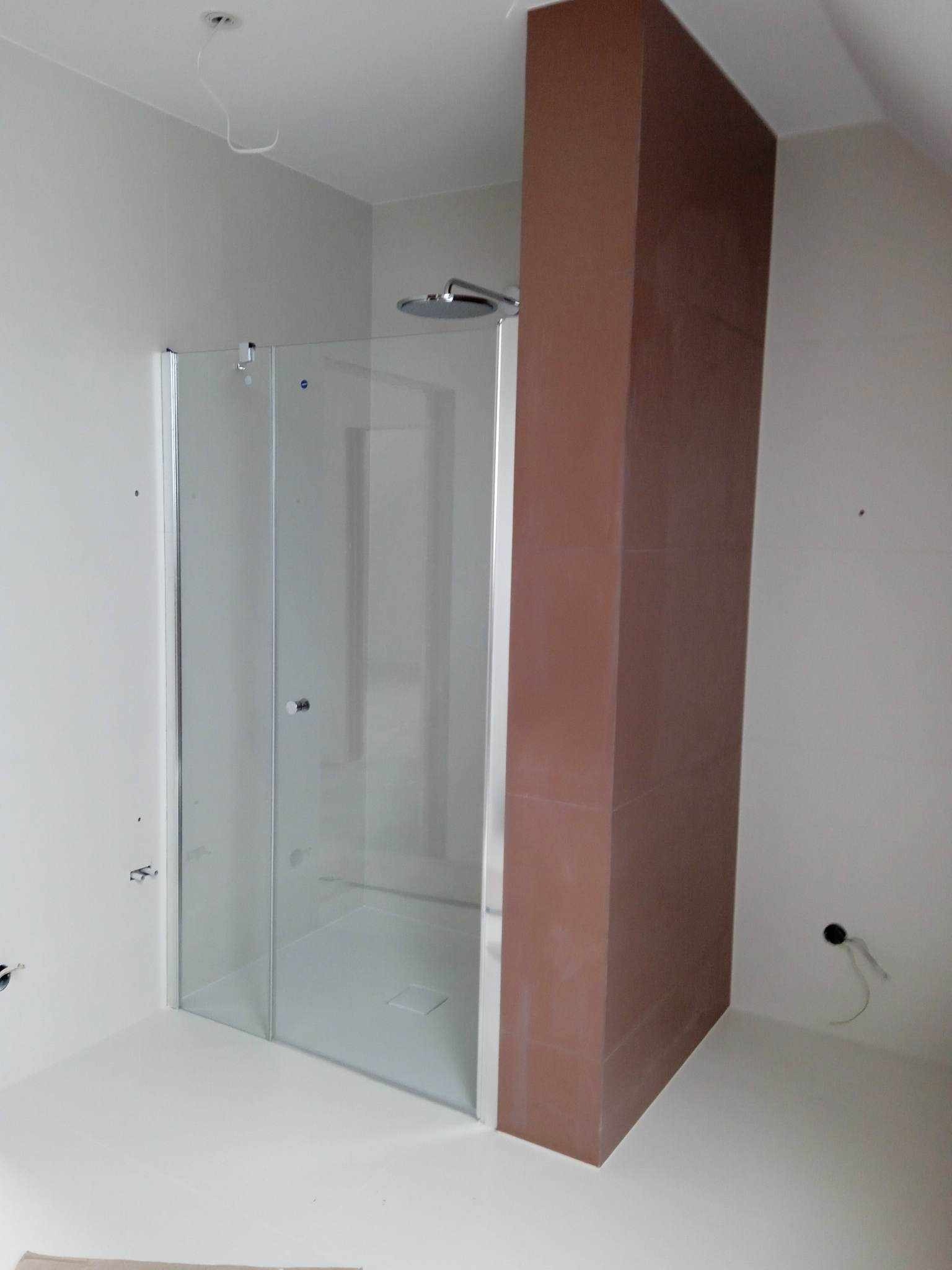 Niedokończona łazienka z kabiną prysznicową ze szklanymi drzwiami i brązową kolumną z płytek, widoczne wystające kable ze ściany i sufitu.