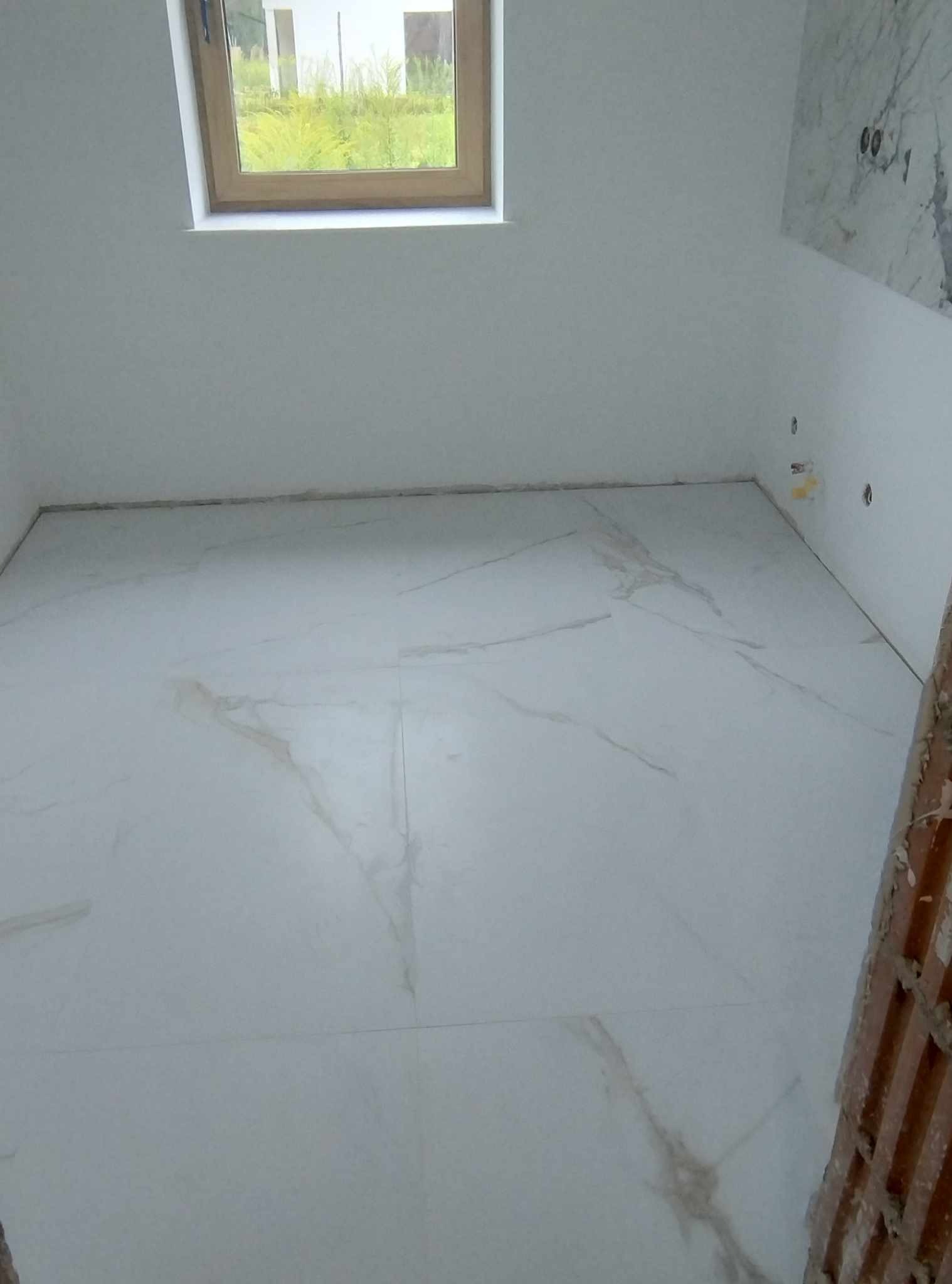 Wnętrze pokoju z nowo ułożoną, jasną podłogą z płytek imitujących marmur, widok na okno i fragment ściany z imitacją marmuru, prace wykończeniowe w toku.