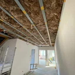 Poddasze w trakcie prac wykończeniowych, widoczne metalowe profile sufitowe z wypełnieniem z wełny mineralnej, drabina i przygotowane płyty izolacyjne oraz częściowo wykończone ściany.