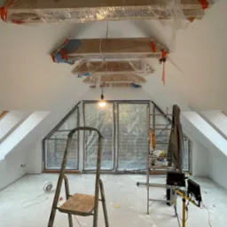 Poddasze w trakcie remontu: świeżo pomalowane białe ściany, zabezpieczone folią drewniane elementy konstrukcyjne sufitu, drabiny, okna dachowe z widokiem na zewnątrz, rozstawione narzędzia i sprzęt...