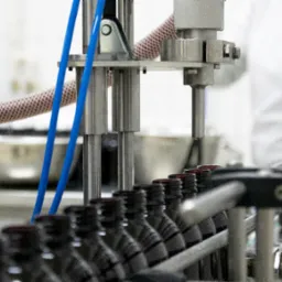 Automatyczna linia produkcyjna z rzędem małych, ciemnych butelek pod dyszą, w tle pracownik w białym fartuchu.