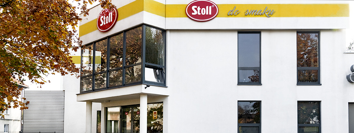 Elewacja budynku z logo Stoll i napisem 'do smaku' nad oknami, jesienne drzewo po lewej stronie.