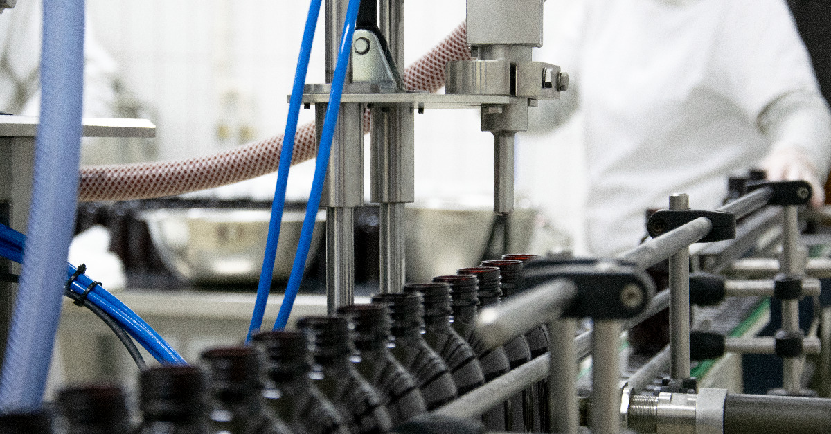 Automatyczna linia produkcyjna z rzędem małych, ciemnych butelek pod dyszą, w tle pracownik w białym fartuchu.