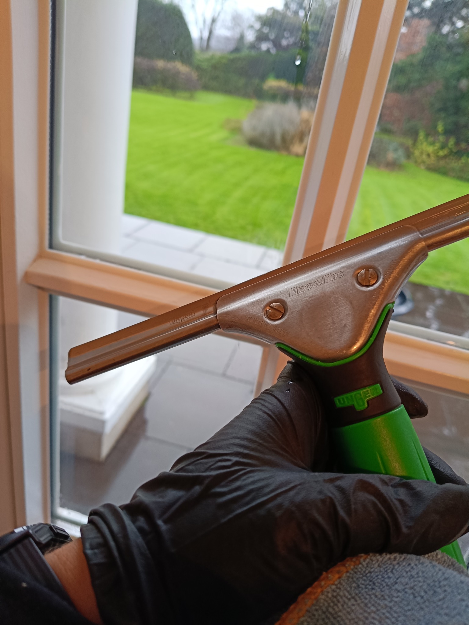 Okno z widokiem na trawnik, czyszczone profesjonalną ściągaczką ErgoTec z zielonym uchwytem, trzymaną w rękawicy ochronnej.