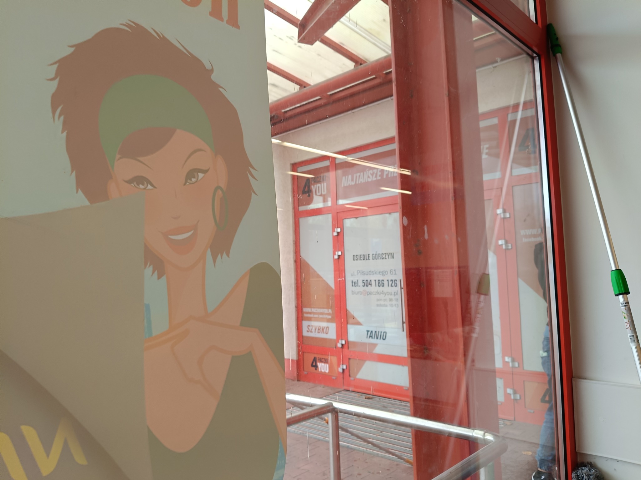 Mop do mycia okien oparty o ramę okienną, widoczne odbicie szyby i plakat z postacią kobiety w tle.