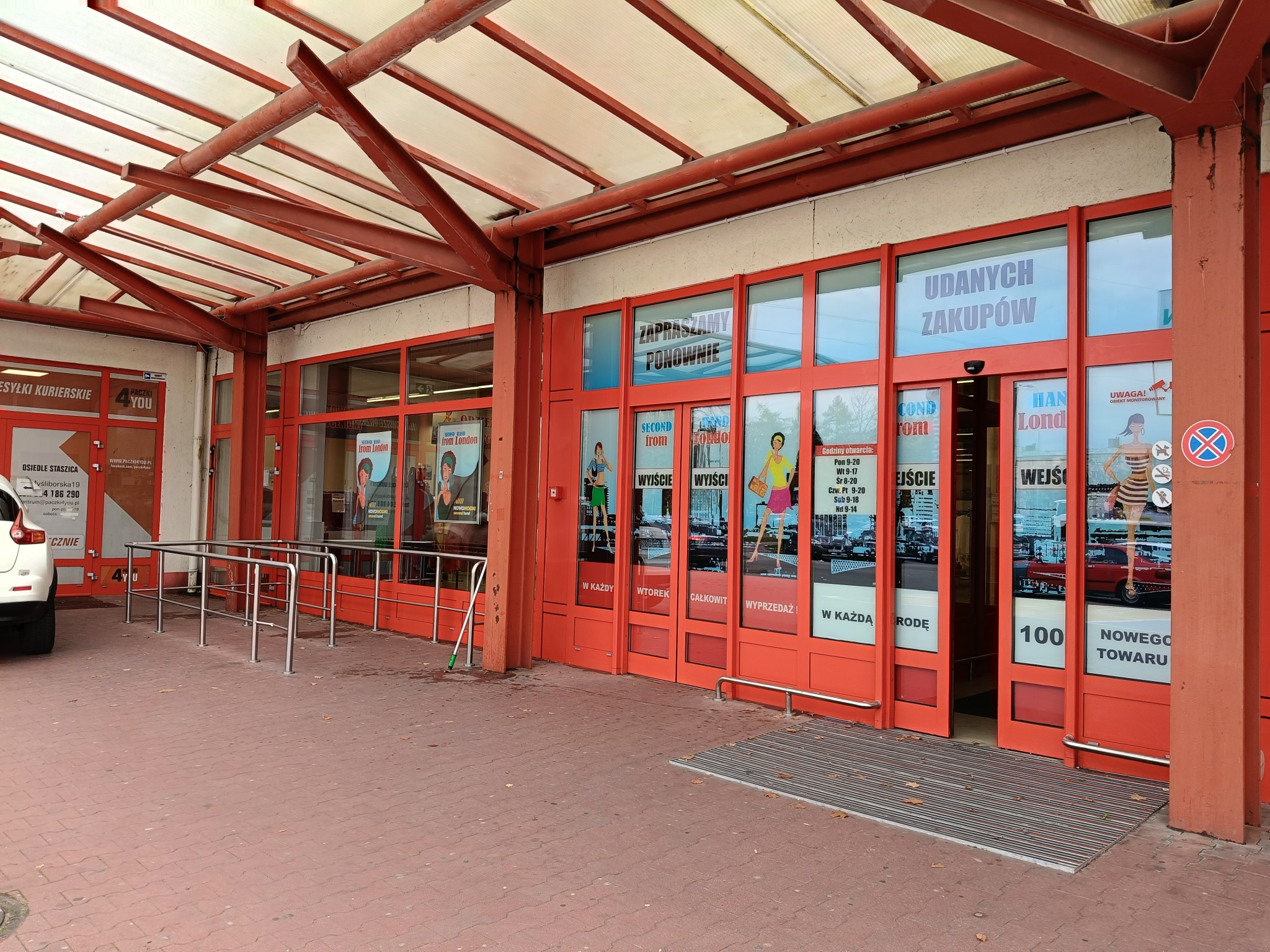 Wejście do sklepu odzieżowego z czerwonymi ramami okiennymi i drzwiami, widoczne plakaty reklamowe i wycieraczka przed wejściem.