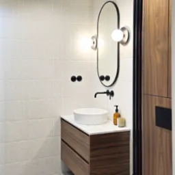 Nowoczesna łazienka z umywalką nablatową na drewnianej szafce, owalnym lustrem z kulistymi lampami, czarną armaturą i grzejnikiem drabinkowym.