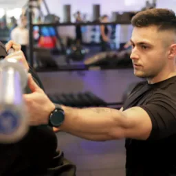 Trener personalny asekuruje klienta podczas wyciskania sztangi na barkach w siłowni, widoczny zegarek na ręce trenera i tatuaż na ramieniu.