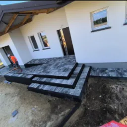 Montaż schodów wejściowych z kostki brukowej do nowoczesnego domu jednorodzinnego z białymi ścianami, widoczny pracownik w pomarańczowej kamizelce i niezagospodarowany teren wokół.