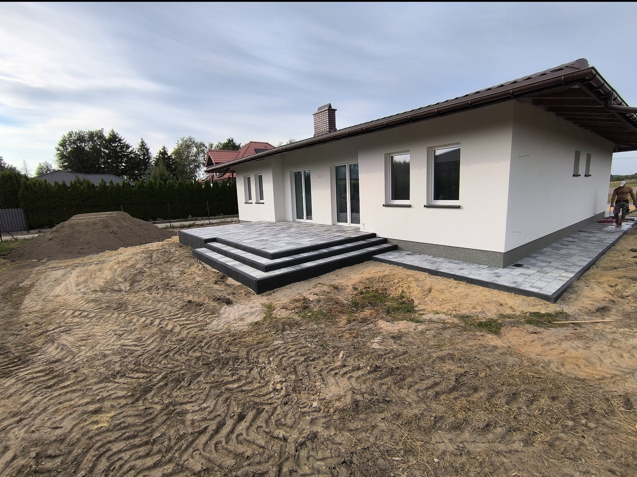 Nowoczesny dom jednorodzinny z białym tynkiem, brązowym dachem i nowo wyłożonym tarasem z szarej kostki brukowej, widoczny stos ziemi i częściowo uporządkowany teren wokół budynku.