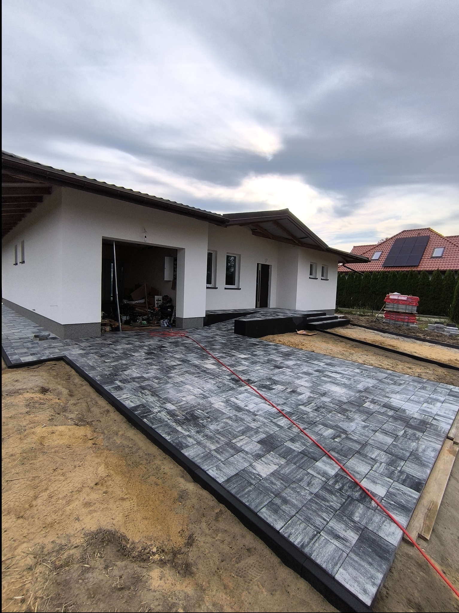 Układanie szarej kostki brukowej przed domem jednorodzinnym, widoczny fragment elewacji, otwarty garaż z narzędziami w środku, oraz ułożone palety z kostką w tle.