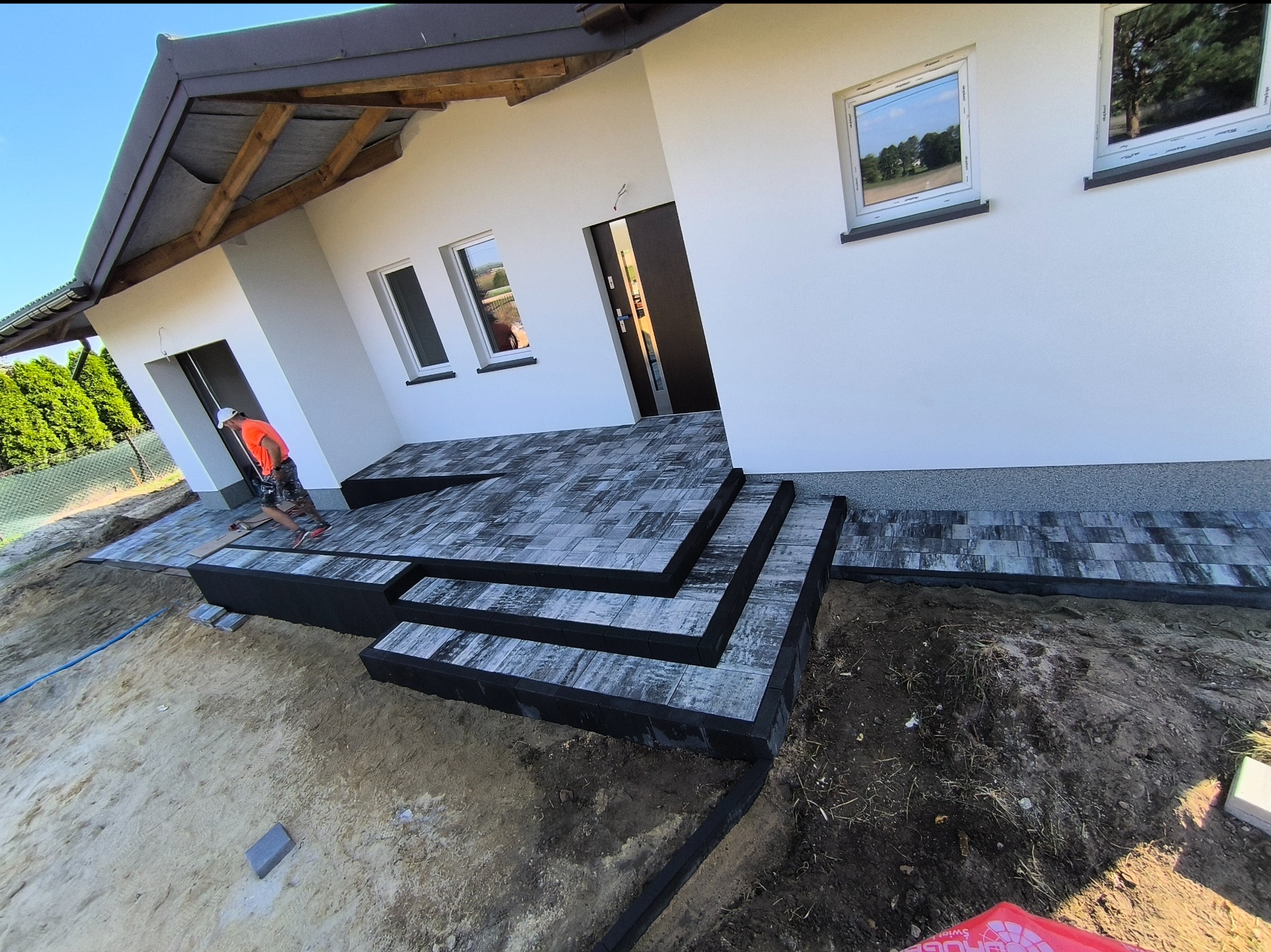 Montaż schodów wejściowych z kostki brukowej do nowoczesnego domu jednorodzinnego z białymi ścianami, widoczny pracownik w pomarańczowej kamizelce i niezagospodarowany teren wokół.