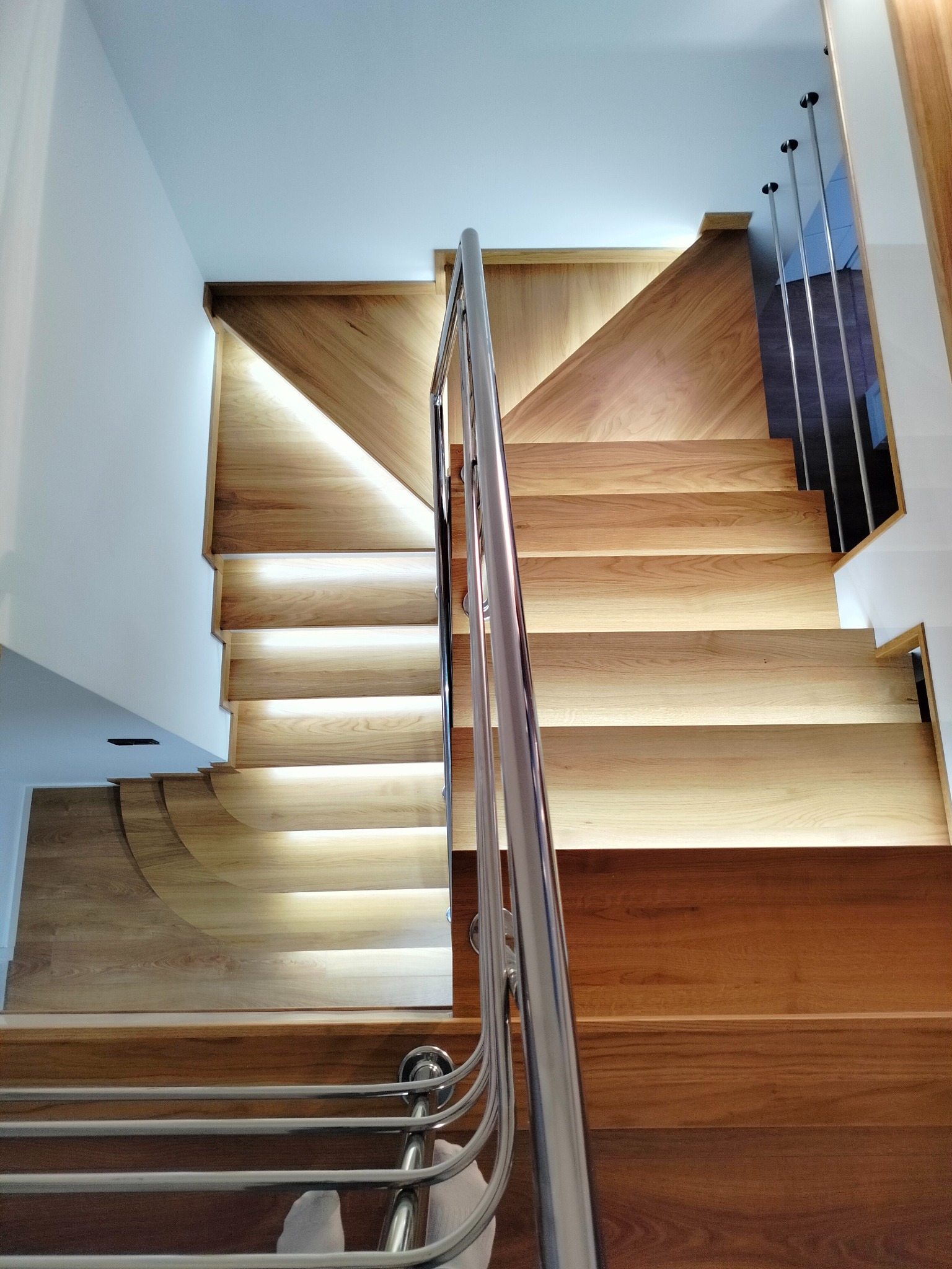 Nowoczesne drewniane schody z podświetleniem LED na każdym stopniu i metalową balustradą, widok z dołu, jasne wnętrze.