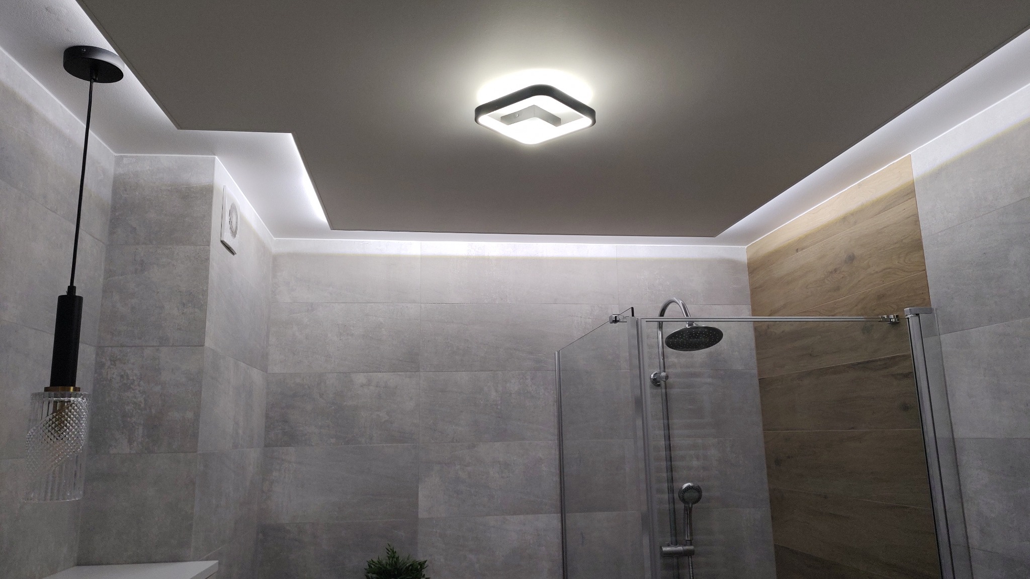 Nowoczesna łazienka z podwieszanym sufitem, oświetleniem LED wzdłuż krawędzi i centralnym kwadratowym plafonem, szklana kabina prysznicowa oraz wisząca lampa z dekoracyjnym kloszem.