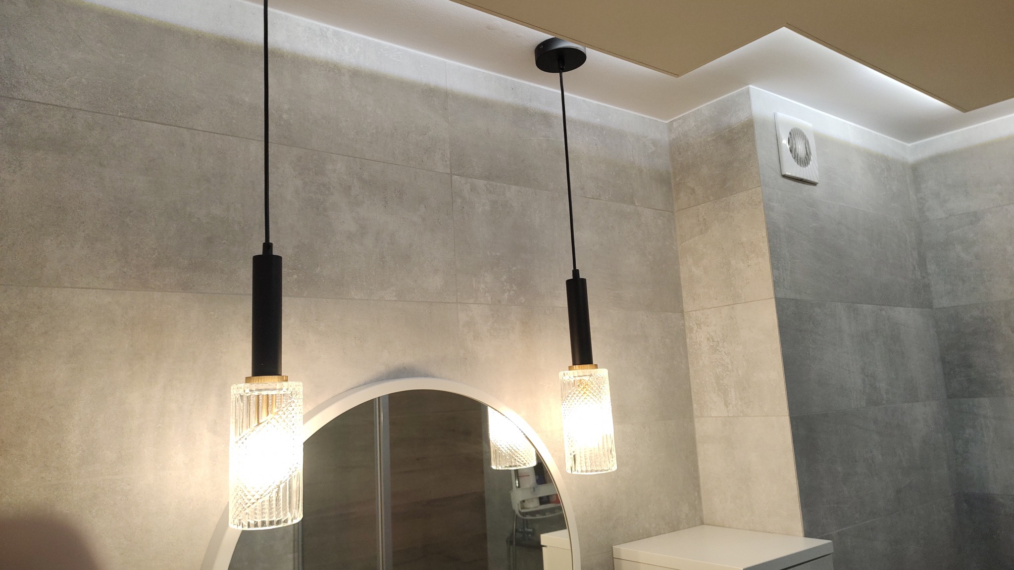 Dwa wiszące, szklane klosze z czarnymi elementami, oświetlające łazienkę z szarymi, matowymi płytkami i okrągłym lustrem.