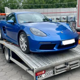 Niebieski sportowy samochód Porsche na metalowej platformie lawety, widok z przodu, w tle zbiorniki i fragment ciężarówki.
