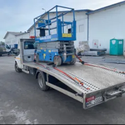 Niebieski podnośnik nożycowy Genie GS-2032 zabezpieczony pomarańczowymi pasami na platformie białej lawety, zaparkowanej na szarym bruku przed budynkiem magazynowym.