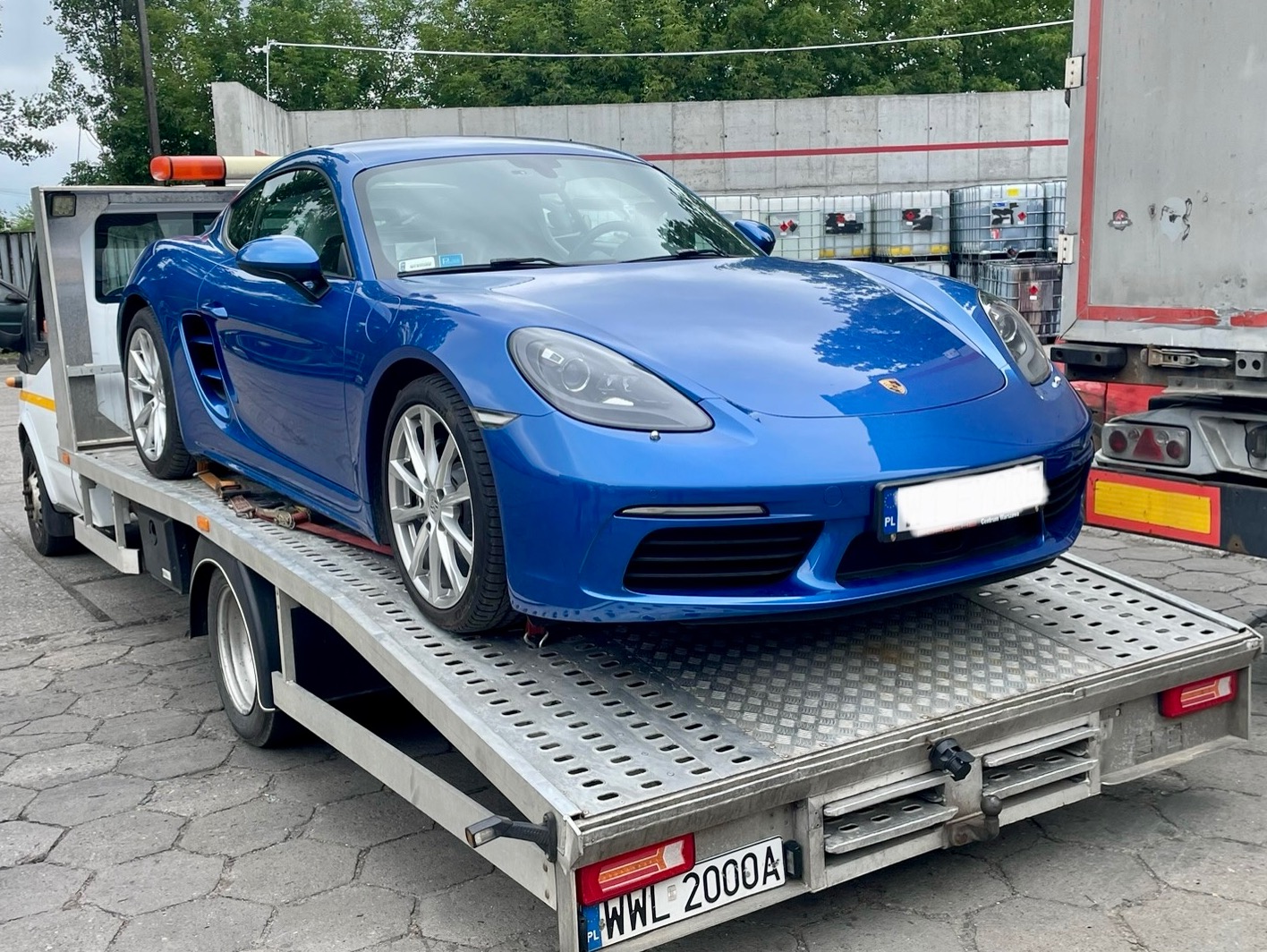 Niebieski sportowy samochód Porsche na metalowej platformie lawety, widok z przodu, w tle zbiorniki i fragment ciężarówki.