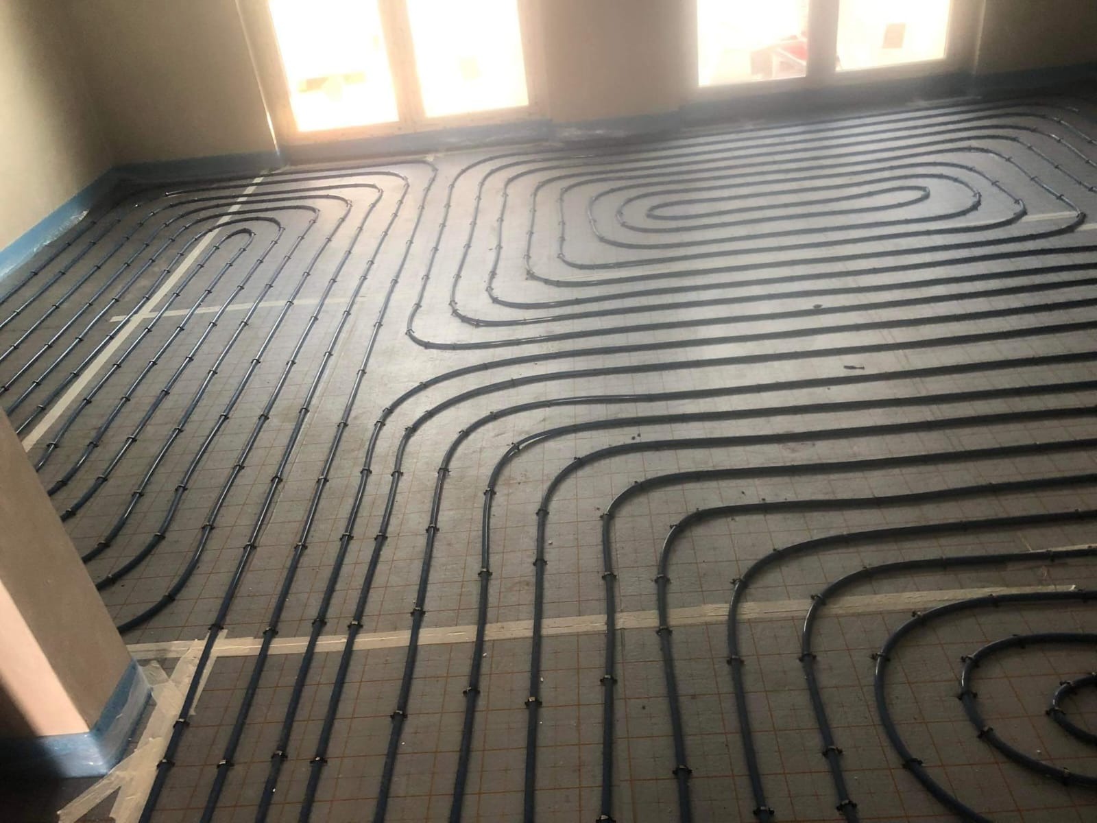 Rozłożone rury ogrzewania podłogowego w kształcie meandrów na podkładzie z siatki, widoczne okno w tle, przygotowane do zalania wylewką.
