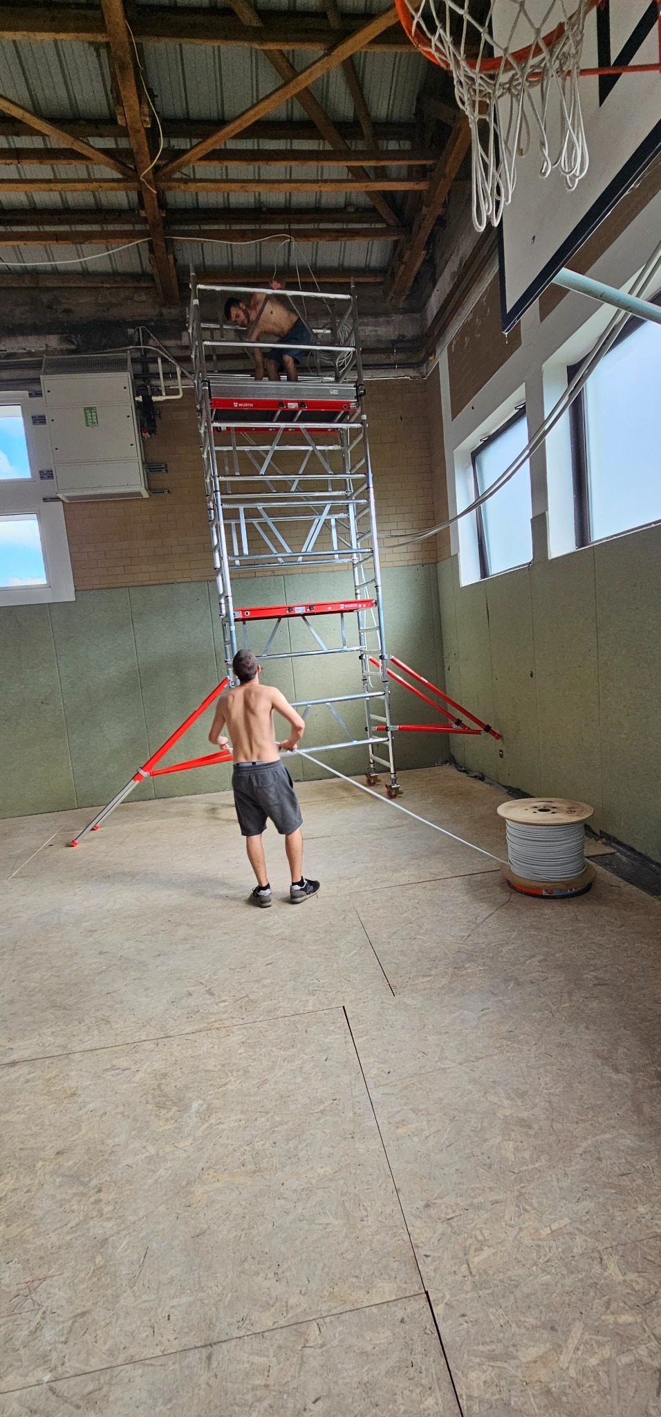 Dwóch mężczyzn bez koszulek na rusztowaniu wewnątrz budynku z odsłoniętym sufitem i instalacją elektryczną, jeden z nich ciągnie kabel z dużej szpuli stojącej na podłodze.