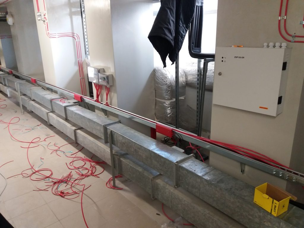 Szare koryta kablowe ułożone wzdłuż ściany w pomieszczeniu technicznym, czerwone przewody, puszki elektryczne, biała skrzynka sterownicza ZSP135-DR i żółte pudełko kartonowe na podłodze.