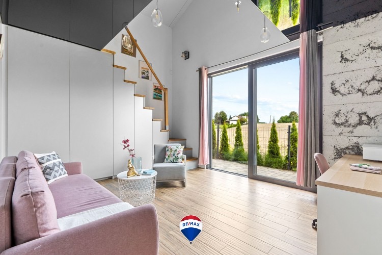Jasne wnętrze domu z drewnianymi schodami, różową sofą i dużym oknem przesuwnym wychodzącym na ogród, podkreślone przez logo RE/MAX na podłodze.