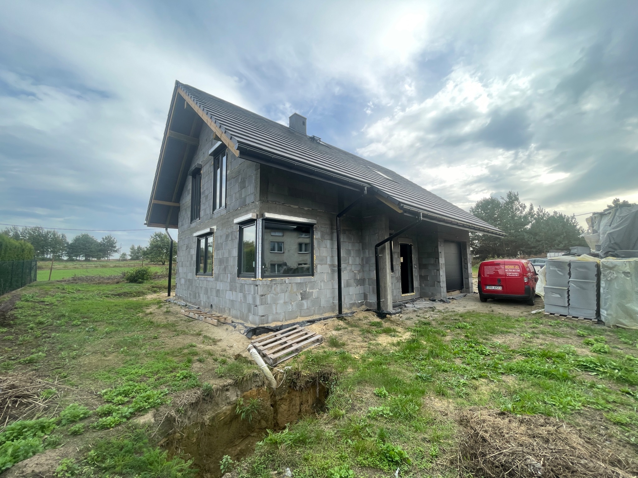 Surowy dom jednorodzinny w trakcie budowy, widoczne szare bloczki, czarny dach z kominem, okna z roletami zewnętrznymi, czerwony samochód dostawczy w tle, wykop ziemny w pierwszym planie.