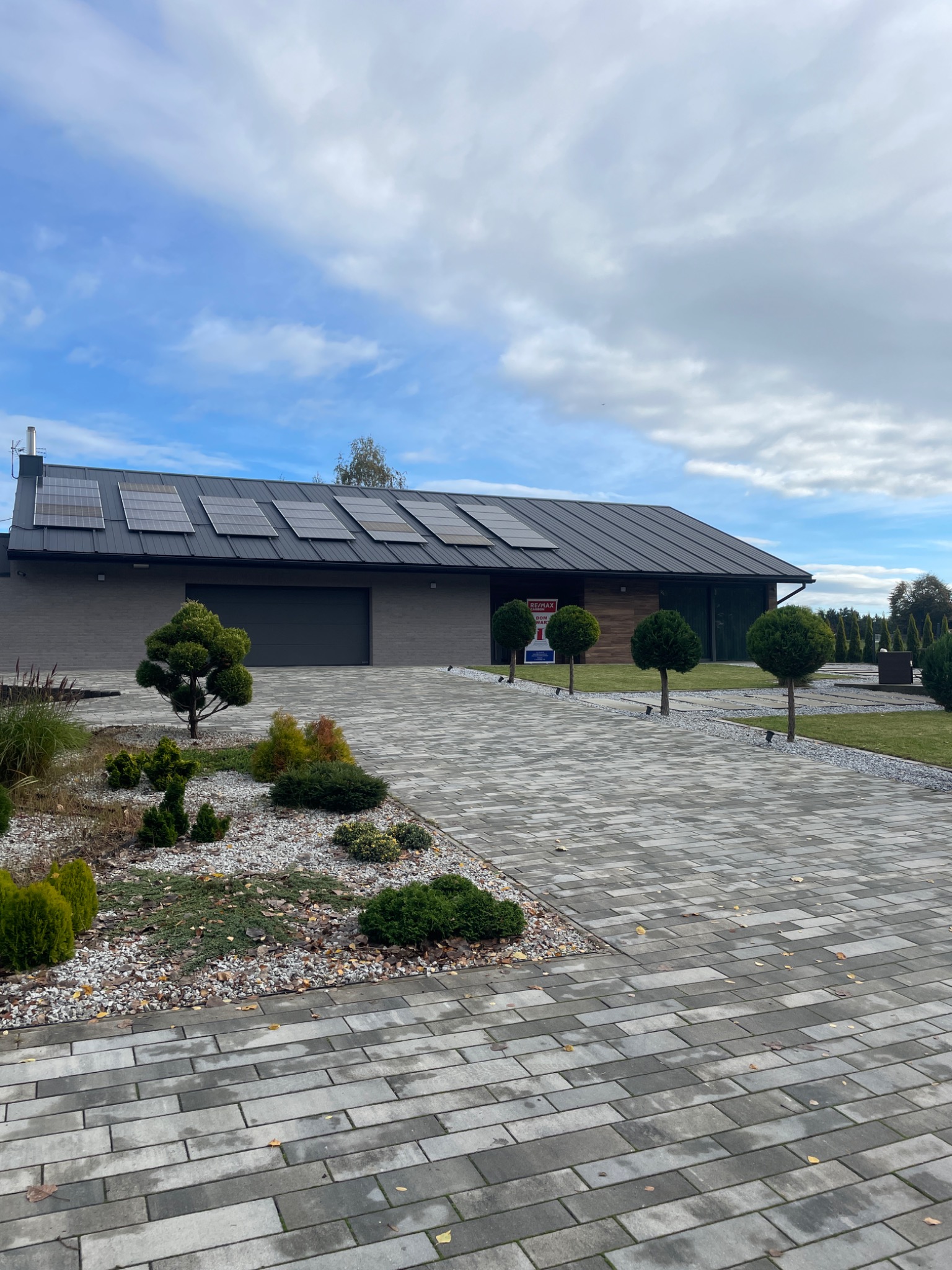 Nowoczesny dom jednorodzinny z panelami słonecznymi na dachu, brukowanym podjazdem i ozdobnymi drzewkami kulistymi, z tabliczką z napisem 'Sprzedam' w oknie.