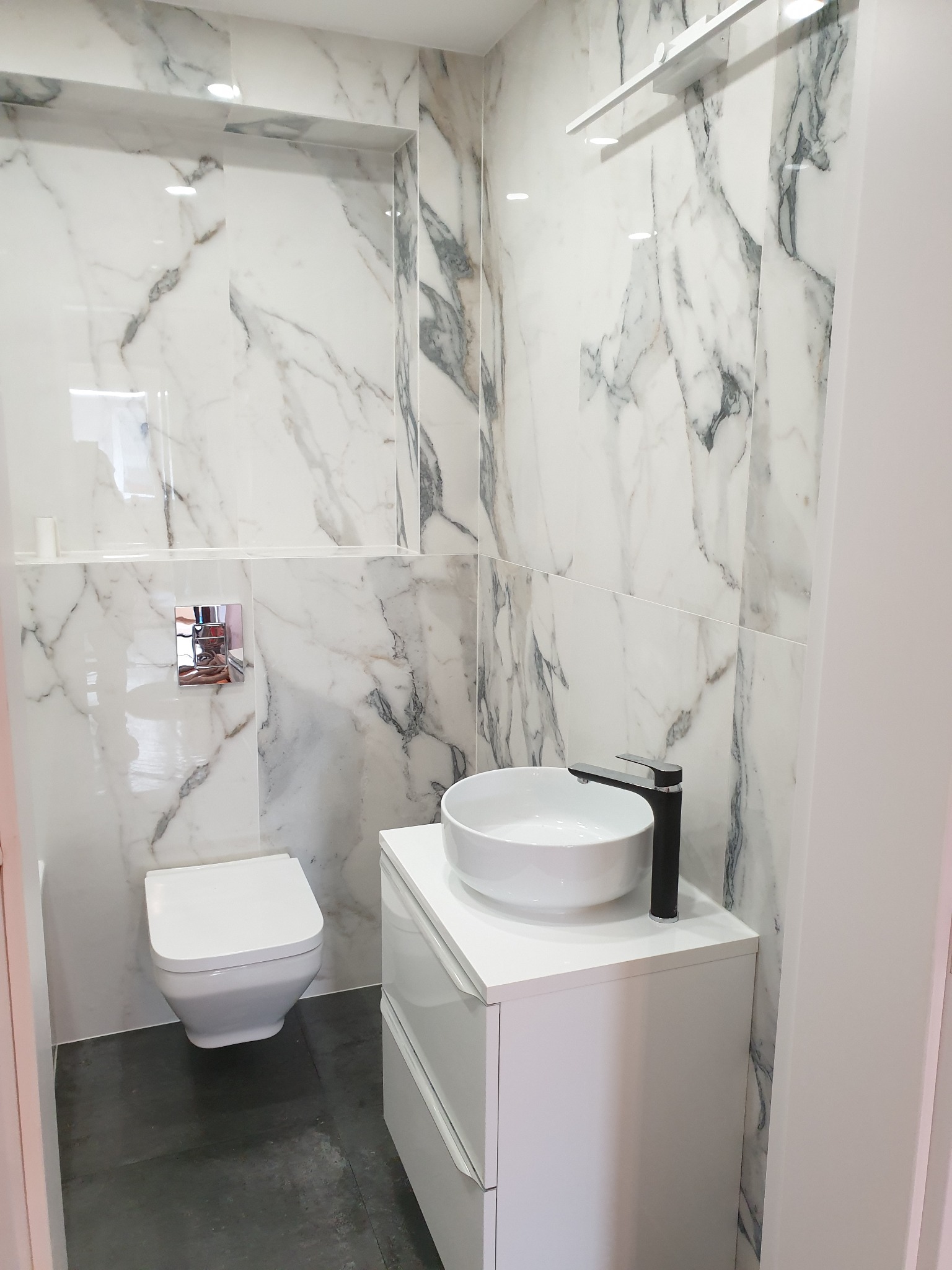 Nowoczesna łazienka z marmurowymi płytkami, wiszącą toaletą, białą umywalką nablatową i czarną armaturą, oświetleniem punktowym w suficie.