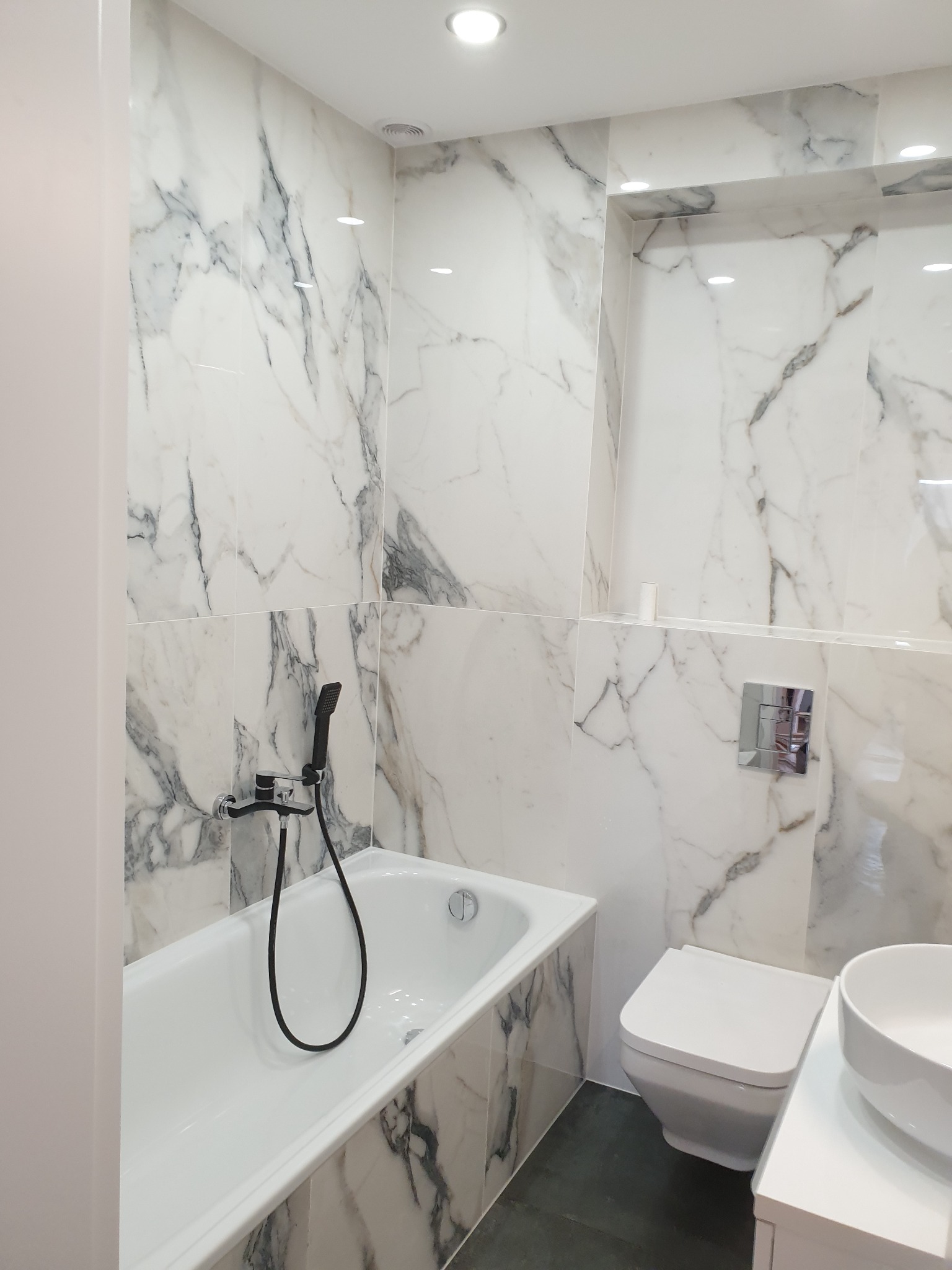 Nowoczesna łazienka wykończona białymi płytkami imitującymi marmur, z wanną, wiszącą toaletą i umywalką nablatową, oświetlona punktowo