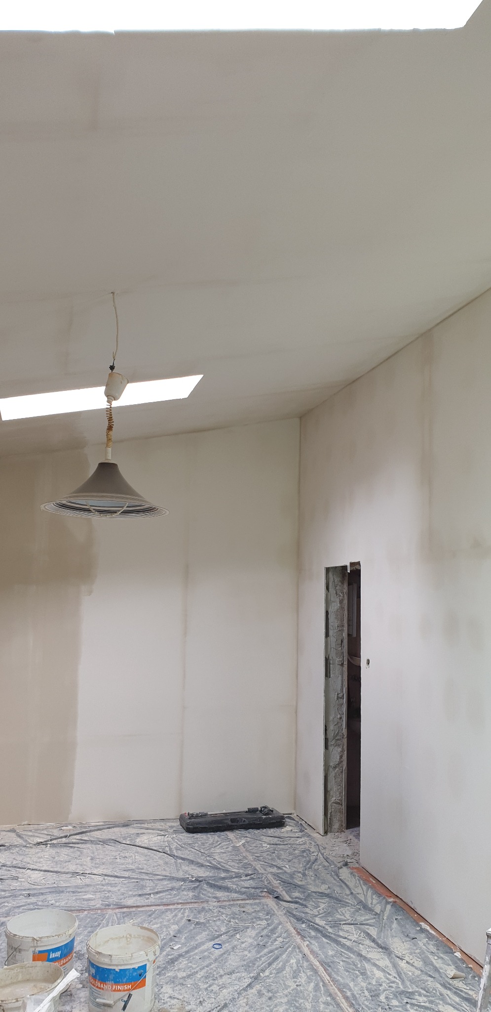 Wnętrze pokoju w trakcie prac wykończeniowych; ściany i sufit po nałożeniu gładzi, podłoga zabezpieczona folią malarską, widoczne wiadra z gładzią i lampa wisząca na kablu.