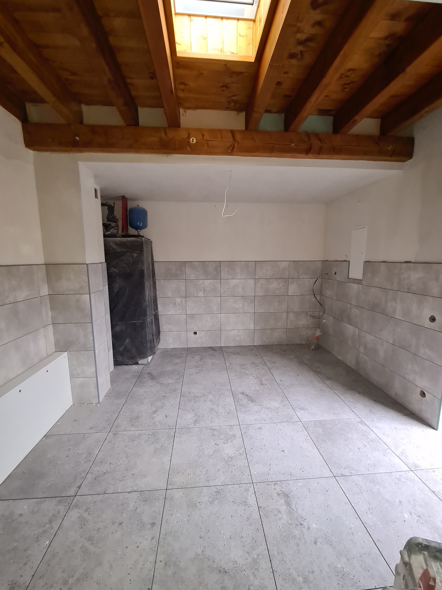 Szare, kwadratowe płytki podłogowe i ścienne w trakcie montażu, widoczne drewniane belki stropowe i okno dachowe, instalacja elektryczna wystająca ze ściany, zbiornik owinięty folią ochronną.