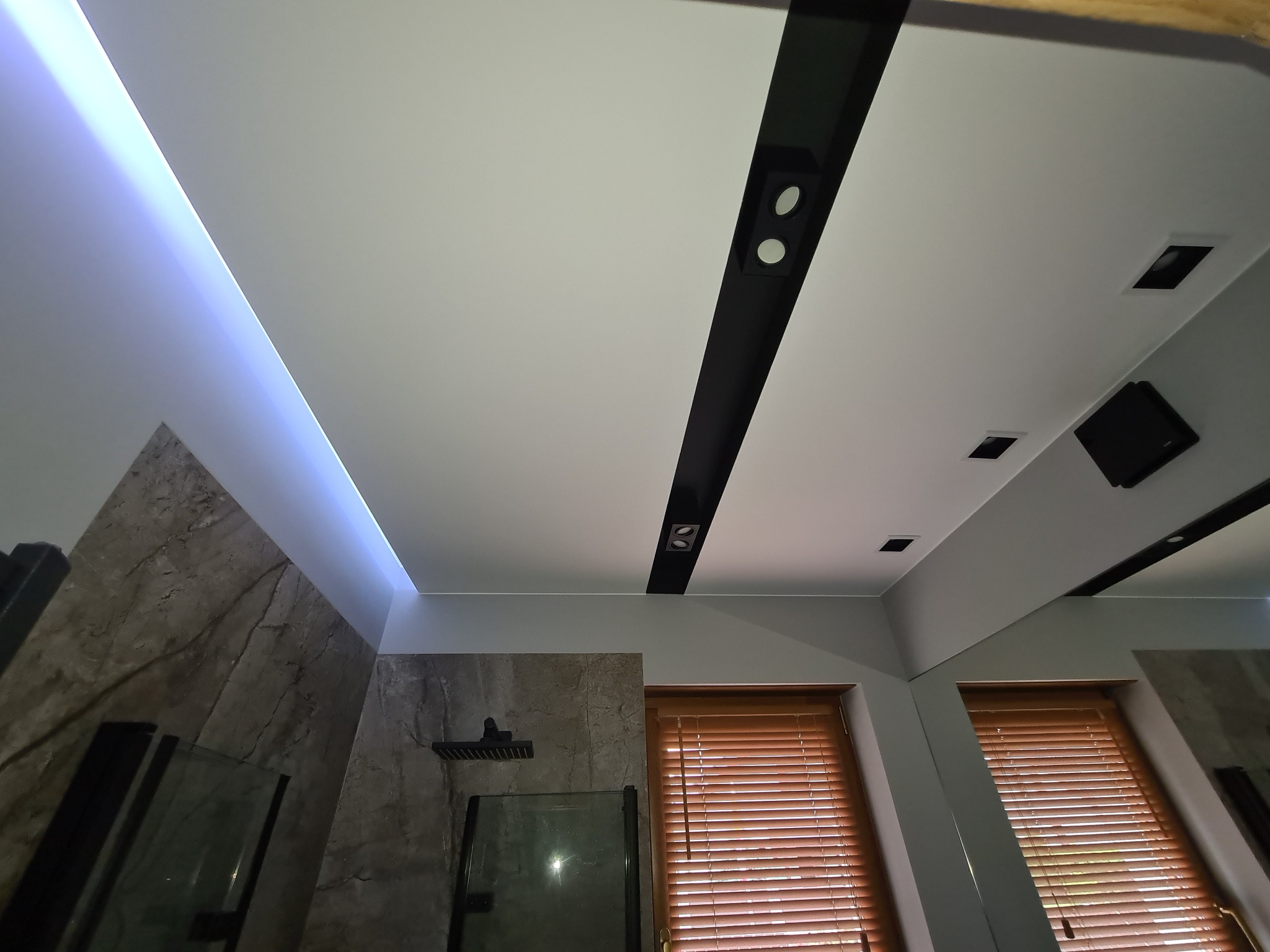 Nowoczesna łazienka z podwieszanym sufitem z oświetleniem LED, czarnymi listwami z punktowymi lampami, marmurowymi płytkami na ścianach i drewnianymi żaluzjami w oknach.