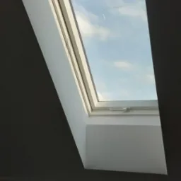 Okno dachowe z widokiem na błękitne niebo z chmurami, zamontowane w szarej ścianie w trakcie wykańczania, widoczne ślady po pracach budowlanych.