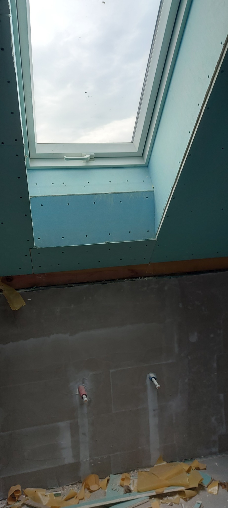 Montaż okna dachowego w ścianie z płyt kartonowo-gipsowych, widoczne szpachlowane ściany i wystające rury instalacji wodnej.