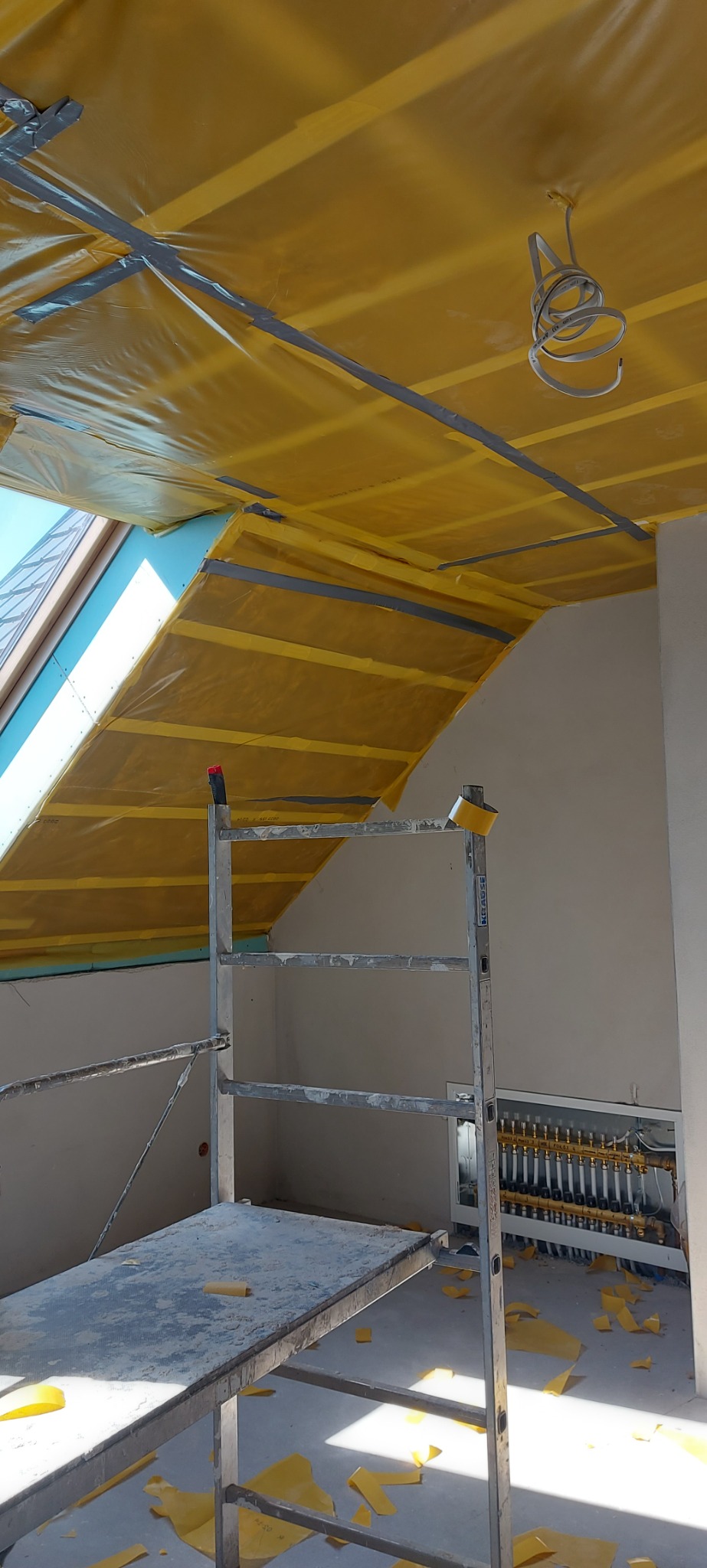 Wnętrze poddasza w trakcie prac wykończeniowych, widoczna żółta folia zabezpieczająca przymocowana taśmą do konstrukcji dachu i sufitu, aluminiowe rusztowanie oraz fragment instalacji grzewczej...