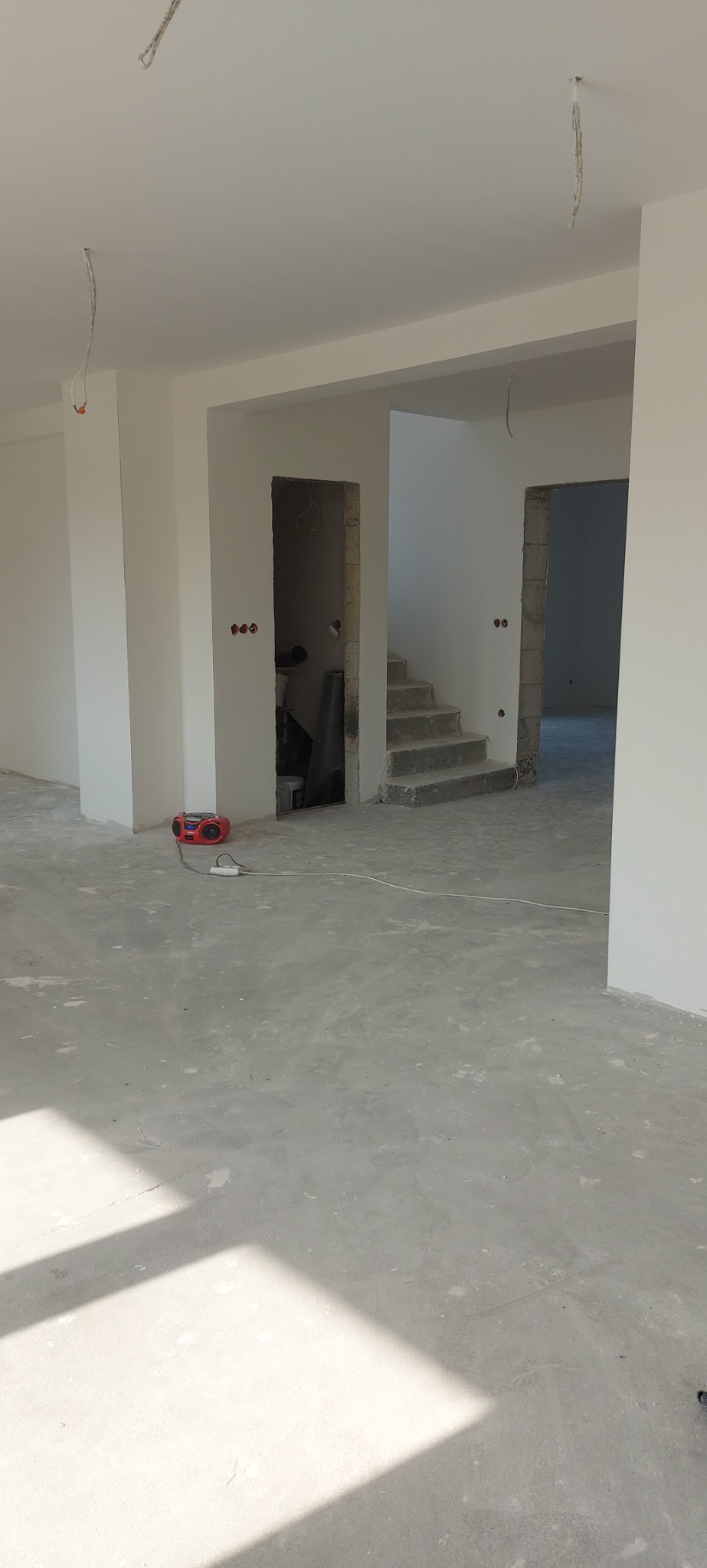 Wnętrze w stanie deweloperskim z surowymi ścianami, betonową posadzką i widocznymi instalacjami elektrycznymi, z czerwonym radiem stojącym na podłodze.