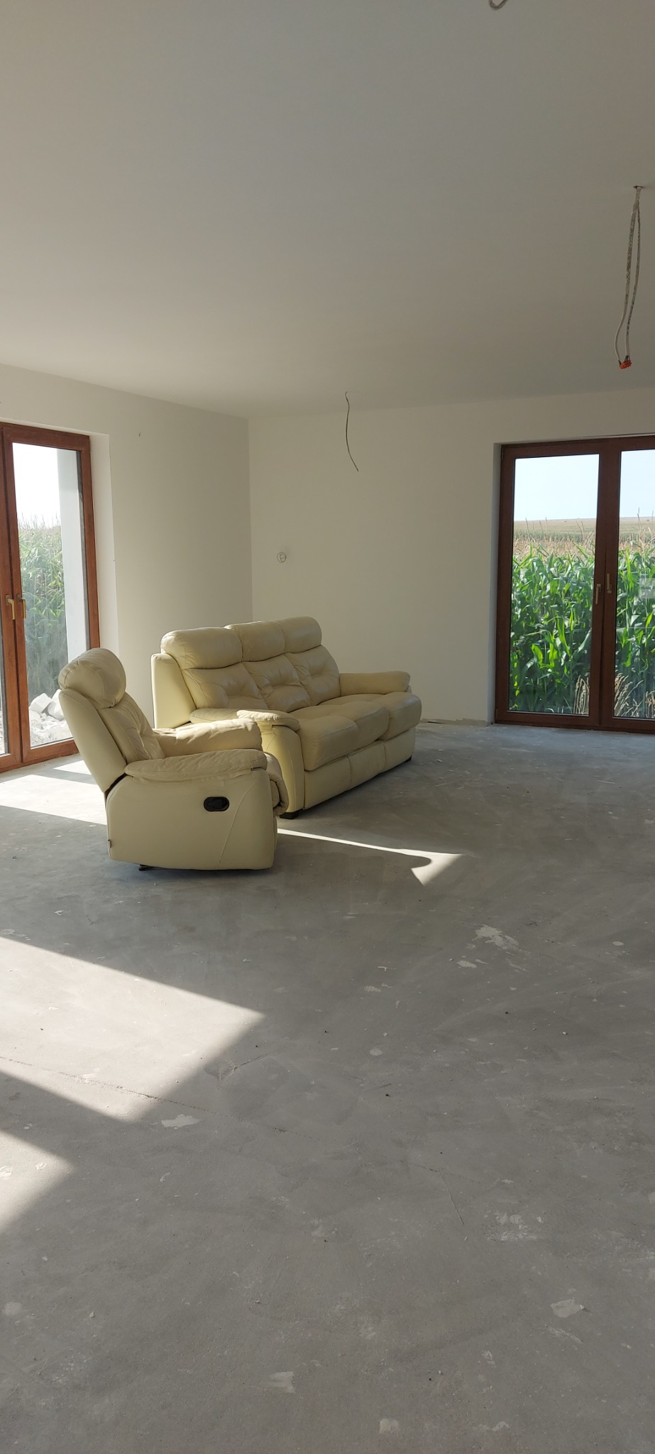 Jasne wnętrze w trakcie wykańczania: kremowa sofa i fotel relaksacyjny na szarym podkładzie podłogowym, widok z okna na pole kukurydzy.