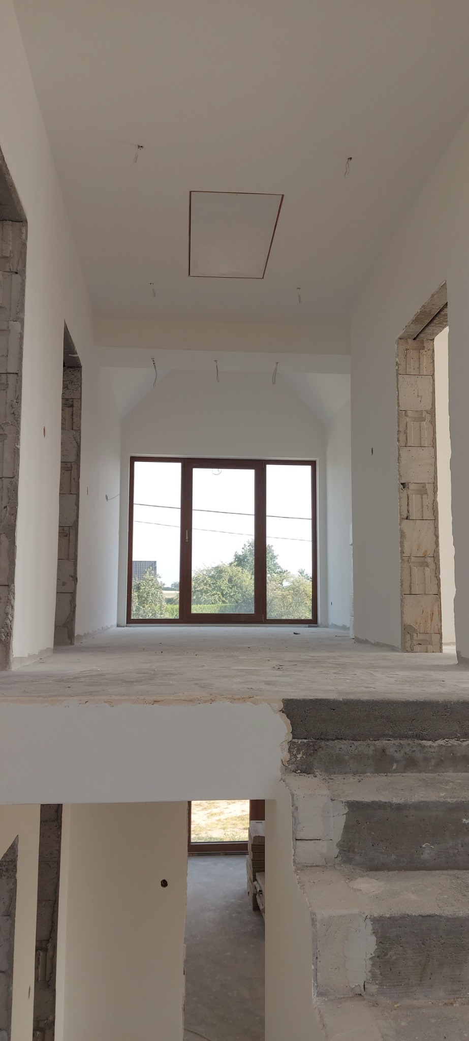 Wnętrze w stanie deweloperskim z widocznymi surowymi ścianami, betonowymi schodami i dużym oknem tarasowym z widokiem na zieleń, elementy instalacji elektrycznej wystają z sufitu.