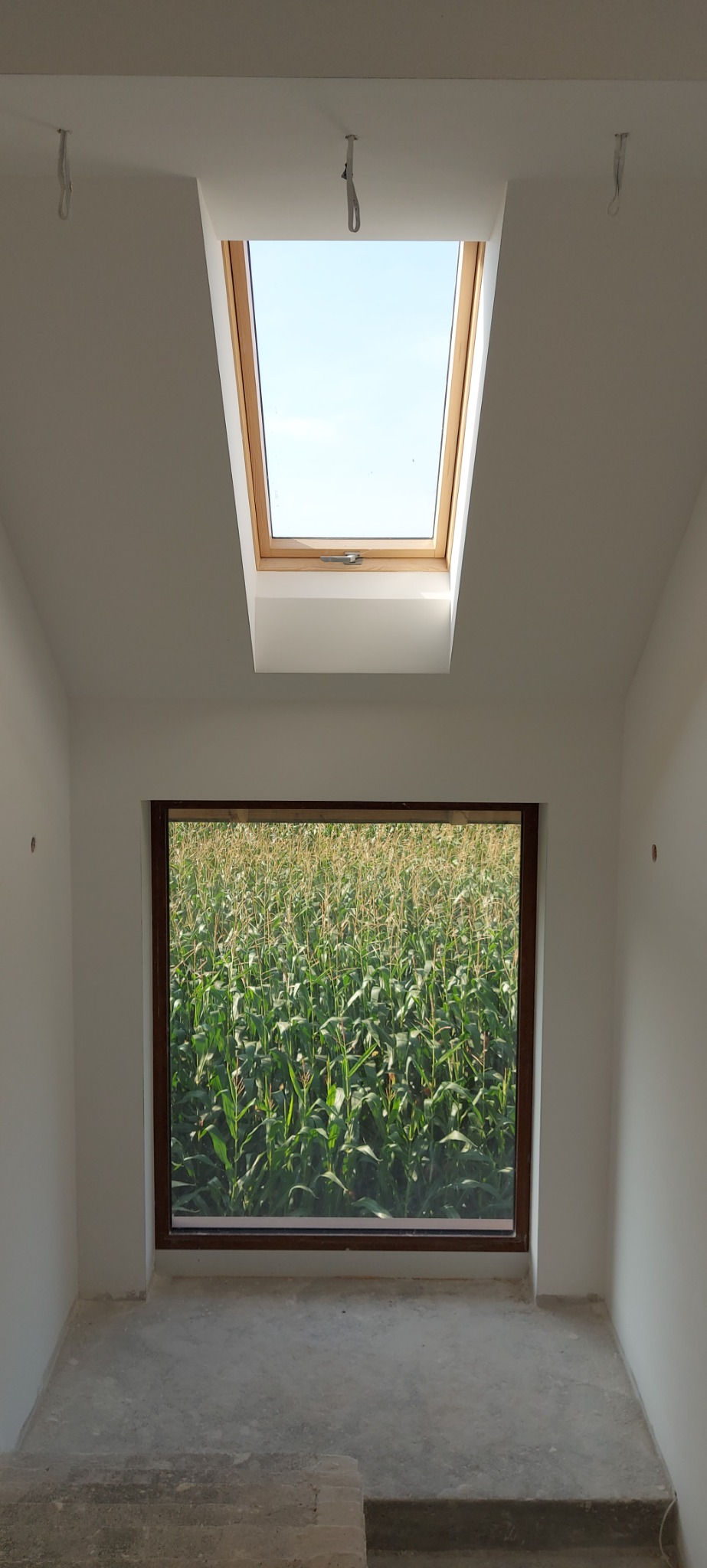 Wnętrze pokoju z dwoma oknami: dachowym i ściennym, z widokiem na pole kukurydzy, ściany pomalowane na biało, surowa podłoga, widoczne uchwyty na suficie.