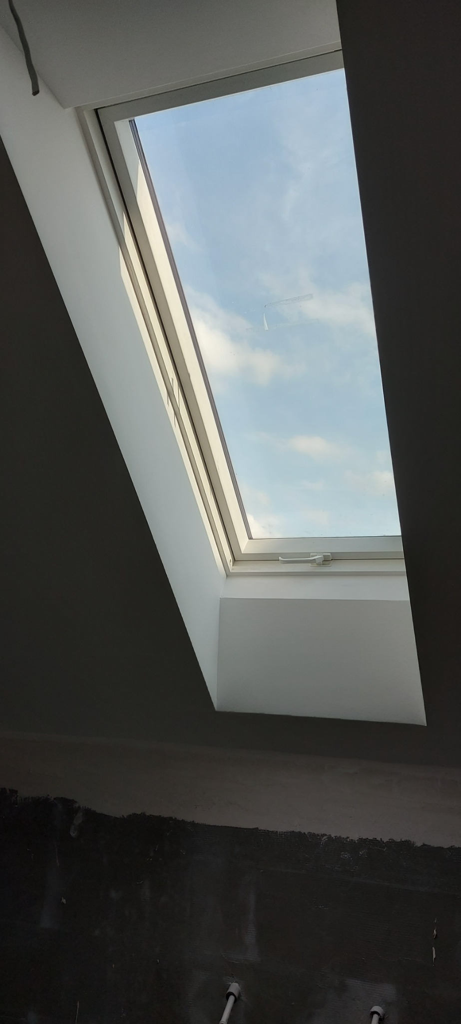 Okno dachowe z widokiem na błękitne niebo z chmurami, zamontowane w szarej ścianie w trakcie wykańczania, widoczne ślady po pracach budowlanych.