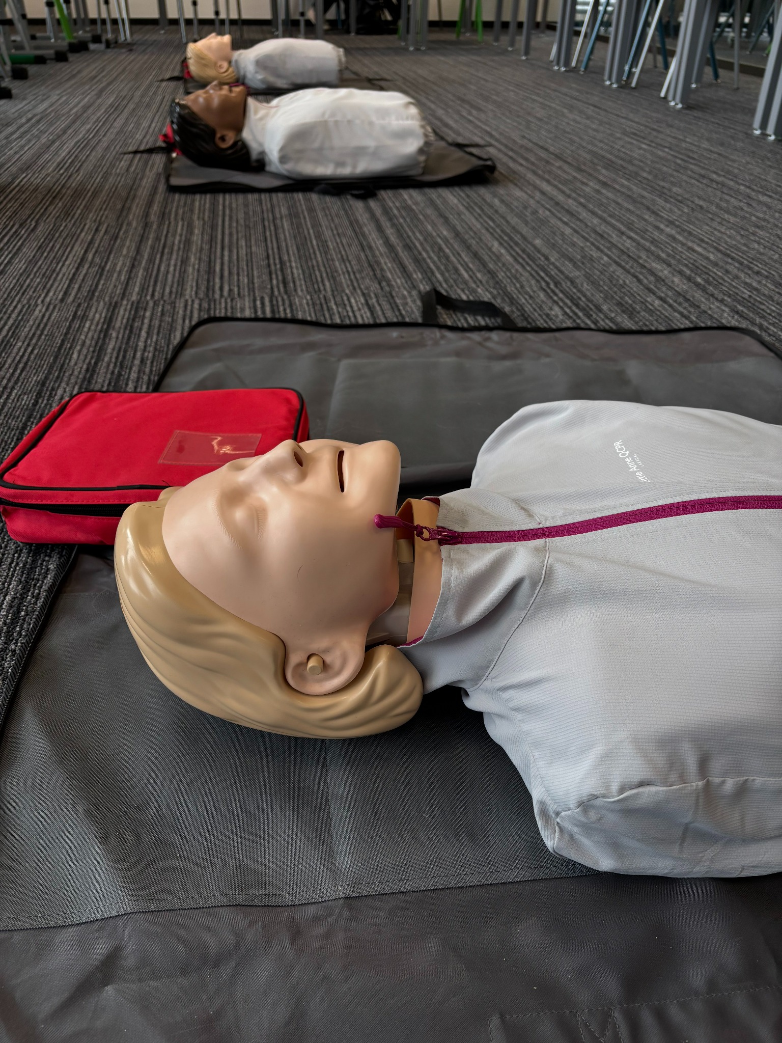 Manekin do nauki RKO z widoczną klatką piersiową i głową, leżący na czarnej macie, obok czerwona apteczka. W tle inne manekiny i krzesła.