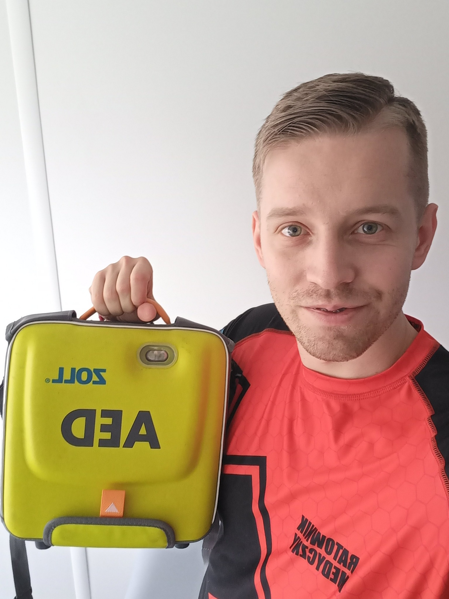 Uśmiechnięty mężczyzna w koszulce ratownika medycznego trzyma żółty defibrylator AED marki Zoll na białym tle.