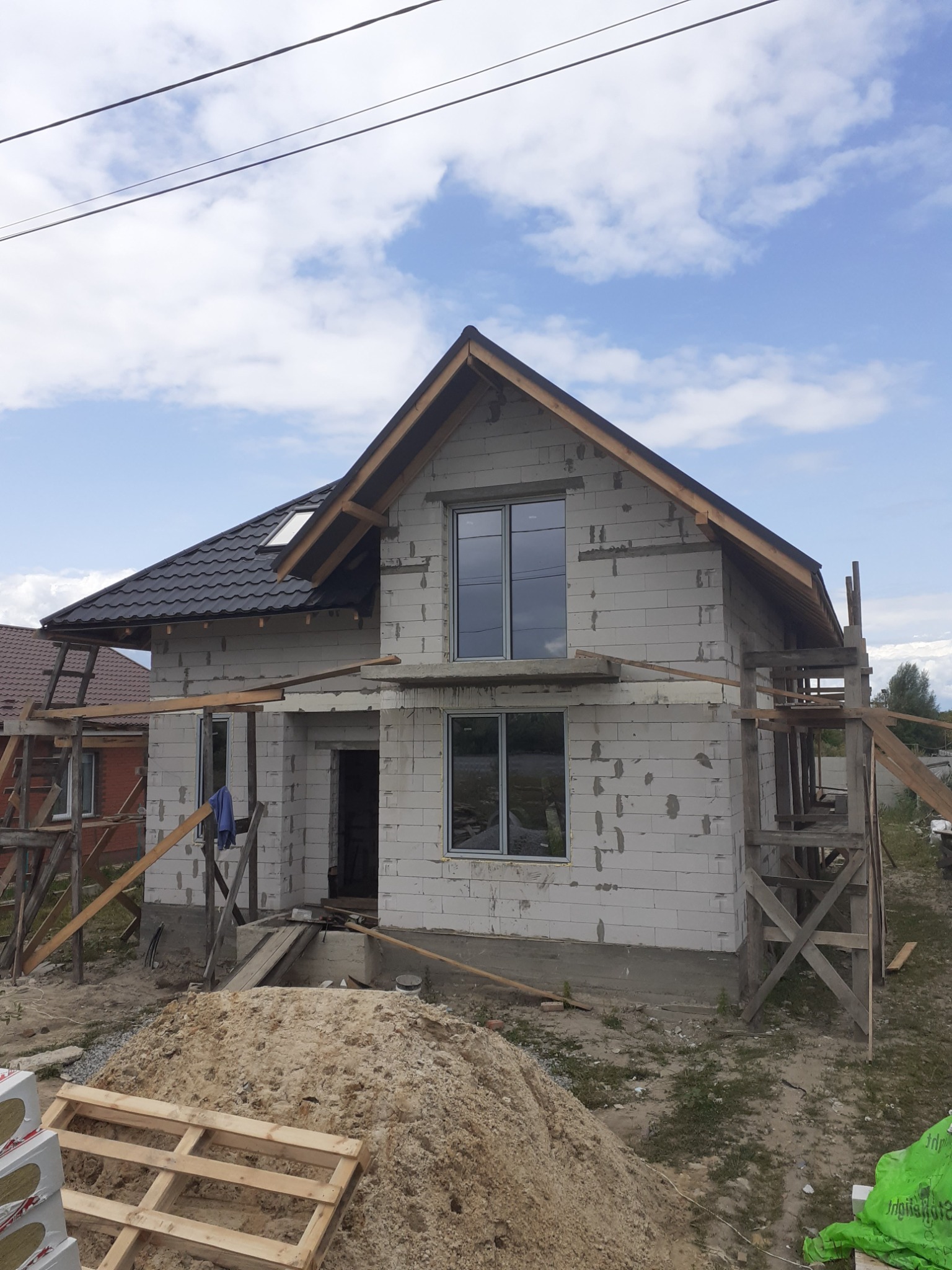 Niedokończony dom z białego bloczka z zamontowanymi oknami i czarnym dachem, widoczne rusztowania i sterta piasku na pierwszym planie.