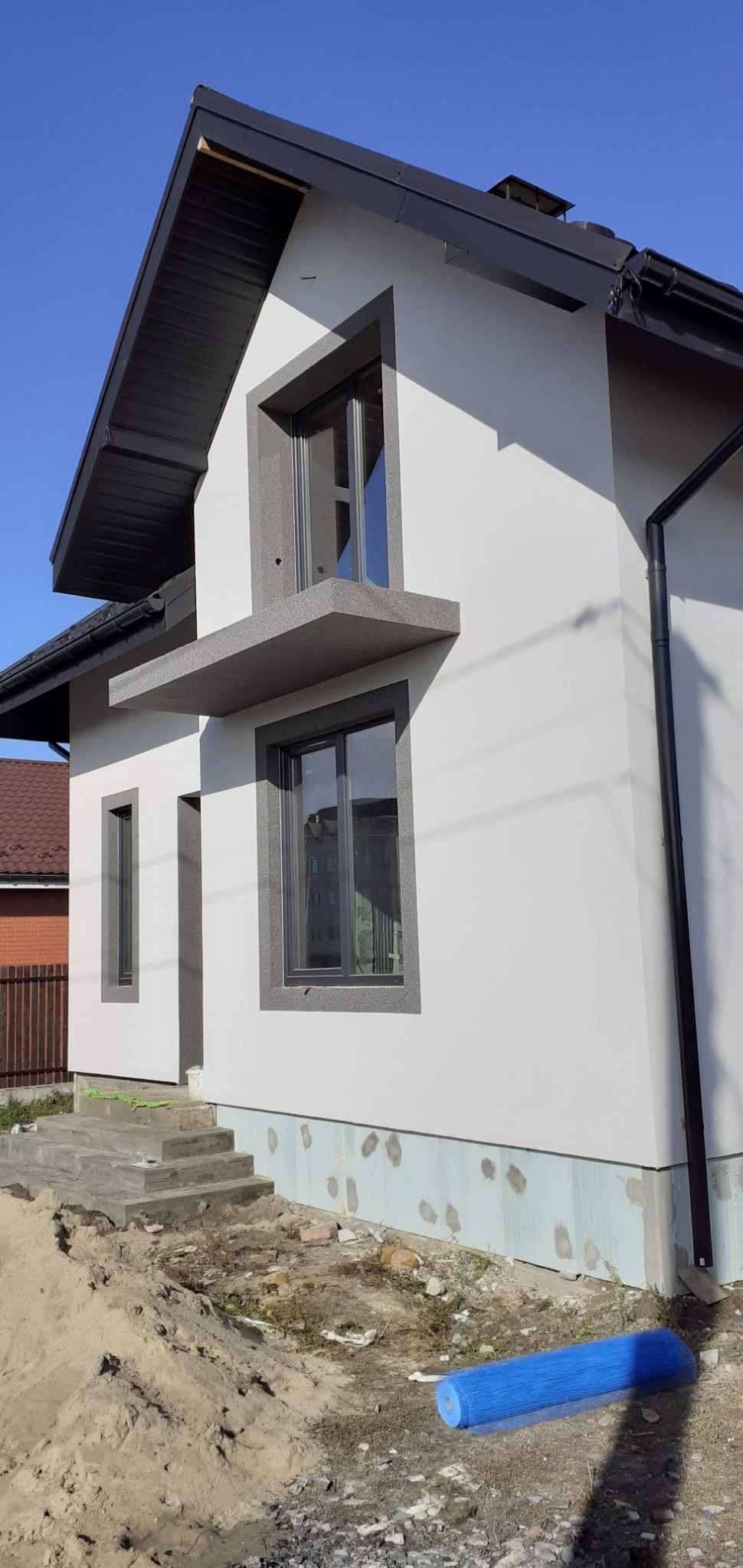 Nowoczesny dom jednorodzinny z ocieploną elewacją w kolorze białym, z szarymi obramowaniami okien i ciemnym dachem, widok z zewnątrz na fragment budynku z widocznymi schodami wejściowymi...