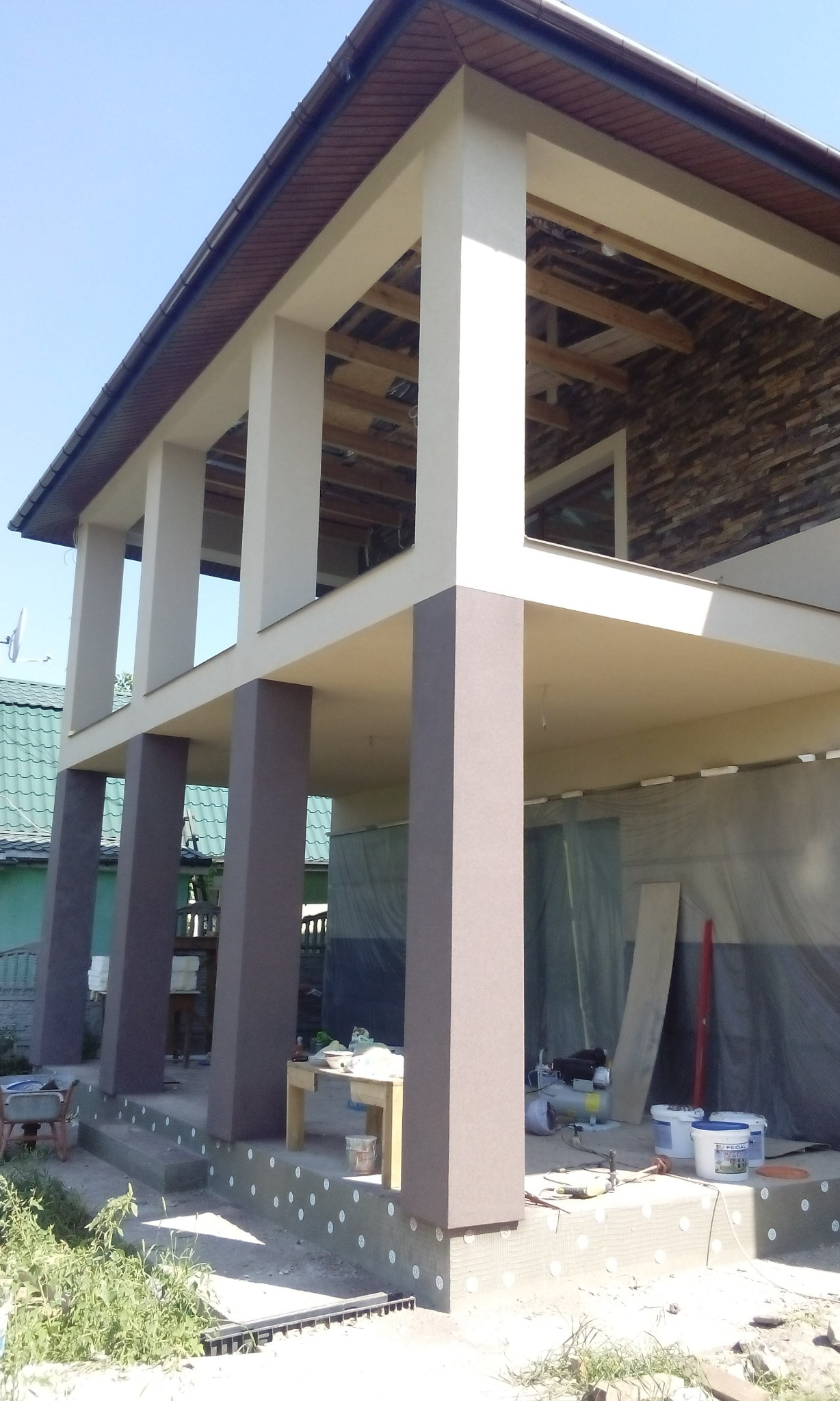 Dom w trakcie ocieplania elewacji z widocznymi kolumnami w dwóch odcieniach brązu i białymi elementami konstrukcyjnymi, styropian na fundamencie, narzędzia i wiaderka z farbą w tle.