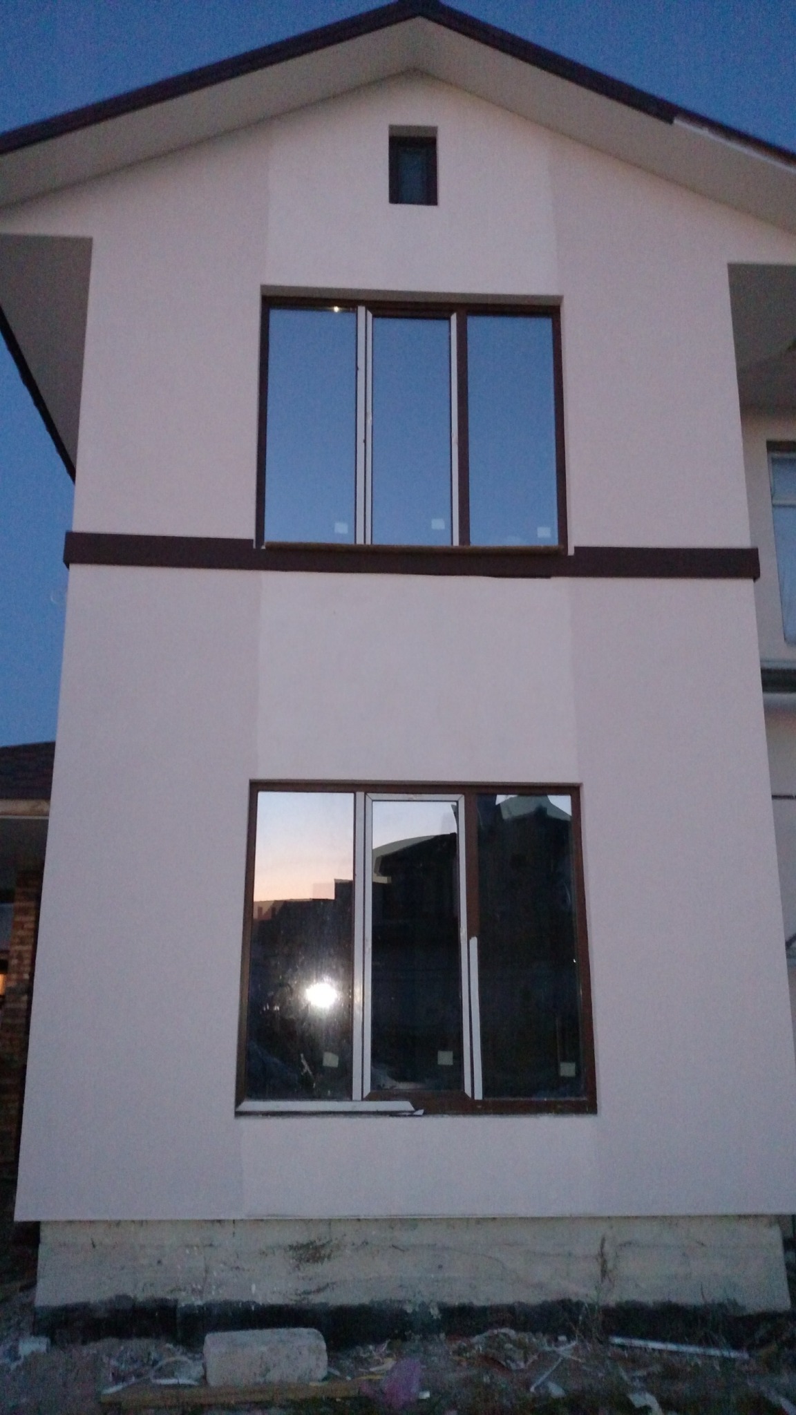 Dwukondygnacyjny dom z ocieploną elewacją w jasnym kolorze, widoczne brązowe ramy okienne i listwa dekoracyjna, teren wokół domu w trakcie prac wykończeniowych.