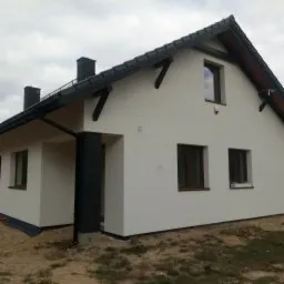 Nowoczesny dom jednorodzinny z białą elewacją, czarnymi elementami dekoracyjnymi z drewna pod dachem i ciemnymi rynnami, widoczny fragment trawnika i niebo z chmurami.