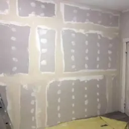 Ściana z płyt kartonowo-gipsowych, wstępnie wyszpachlowana, widoczne łączenia płyt i wkręty, drabina aluminiowa po lewej stronie i zabezpieczona podłoga folią malarską w rogu pokoju.